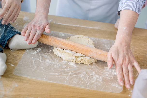 Sheeting dough. Women's hands are holding rolling pin and floured. Hands baking dough with rolling pin. Baking in kitchen. Chef at work. Women at kitchen, little feet of baby on the table - Фото, изображение