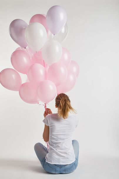 Achteraanzicht van meisje in casual kleding zitten en houden van roze lucht ballonnen op wit - Foto, afbeelding