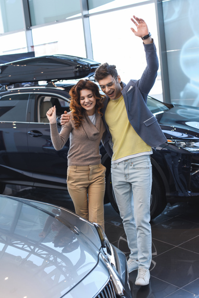 gelukkige man knuffelen krullend aantrekkelijke vrouw, terwijl het vieren van triomf in de auto showroom  - Foto, afbeelding