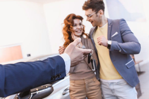 selective focus of car dealer holding keys near cheerful man and woman in car showroom  - Photo, Image
