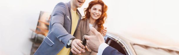 panoramic shot of car dealer giving keys to cheerful man and woman in car showroom  - Photo, Image