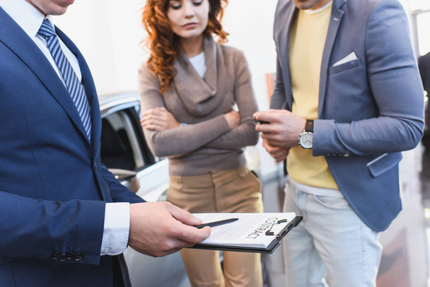 cropped view of car dealer holding clipboard with contract near man and woman with crossed arms  - Fotoğraf, Görsel