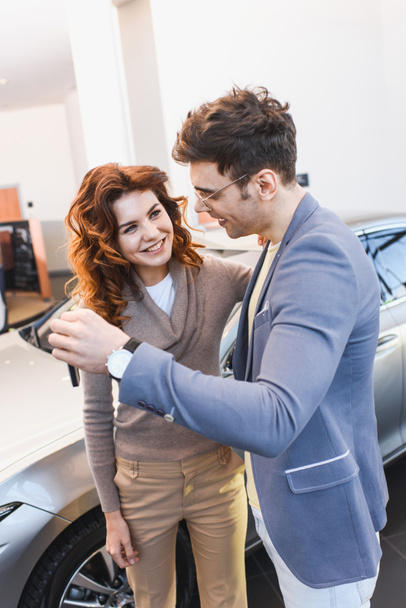 szczęśliwy człowiek w okularach stojących z wesołą kręcone kobiety i trzymając klucze w salonie samochodowym  - Zdjęcie, obraz