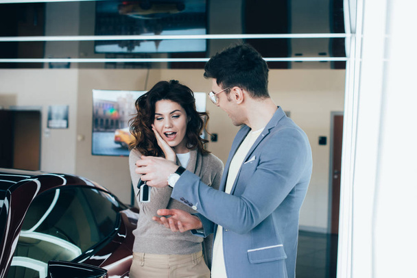cheerful man holding keys near surprised curly girl while standing near red automobile - Foto, Imagen