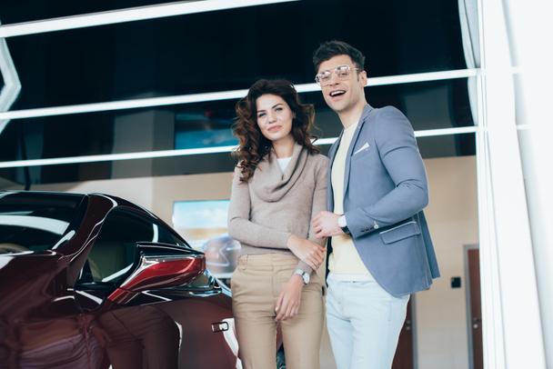 happy woman and handsome man looking at camera while standing near red automobile - Photo, Image