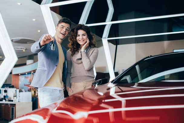 fröhlicher Mann, der Schlüssel in der Hand hält, während er mit einer lächelnden, lockigen Frau neben einem roten Auto steht - Foto, Bild