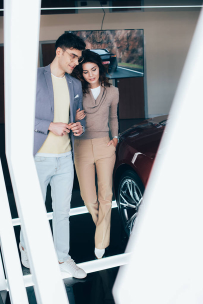селективный фокус стильного мужчины в очках, гуляющего с кудрявой девушкой возле красного автомобиля
 - Фото, изображение