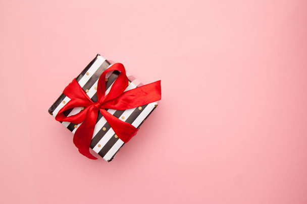Gift or present box in white and black stripes with a red ribbon bow isolated on pink table top view. Minimal flat lay composition for sales, birthday, mother day or christmas. copyspace - Foto, imagen