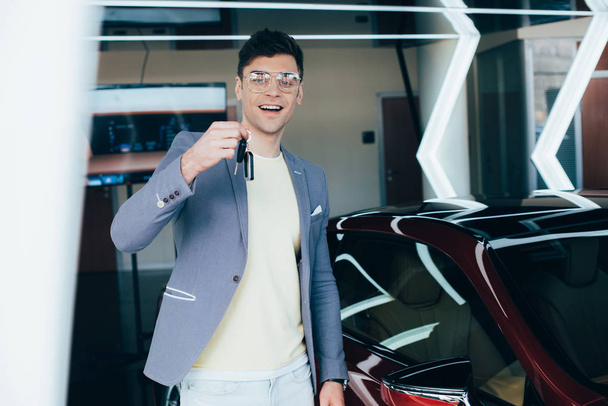 vrolijke man in glazen Holding toetsen terwijl staande in de buurt van rode auto  - Foto, afbeelding
