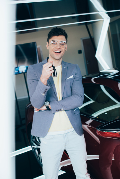 selective focus of happy stylish man in glasses holding keys while standing near red automobile  - Photo, Image