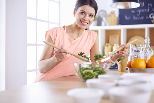 Smiling young woman mixing fresh salad in the kitchen. - Фото, изображение
