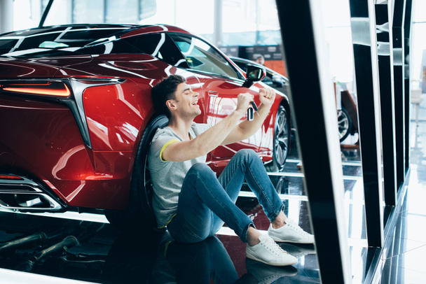 messa a fuoco selettiva di uomo felice seduto sul pavimento vicino alla nuova auto rossa e mostrando sì gesto
 - Foto, immagini