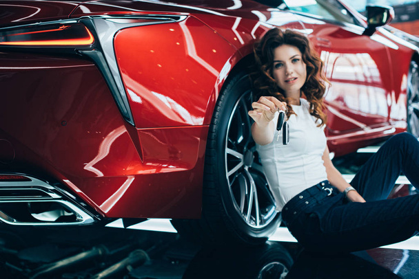 Glückliche Frau sitzt auf dem Boden neben neuem roten Auto und hält Schlüssel in der Hand - Foto, Bild