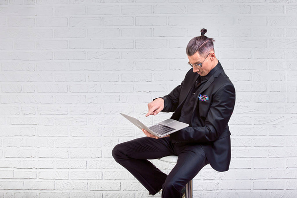 Handsome business man in glasses and suit holding laptop in hands and writing something. Side view. On white brick background - Fotografie, Obrázek