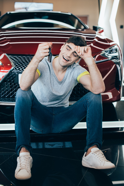 happy handsome man sitting on floor near new red car and holding keys - Fotó, kép