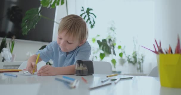 Smiling boy in blue shirt draws on paper with a pencil while sitting at the table in the living room - Video