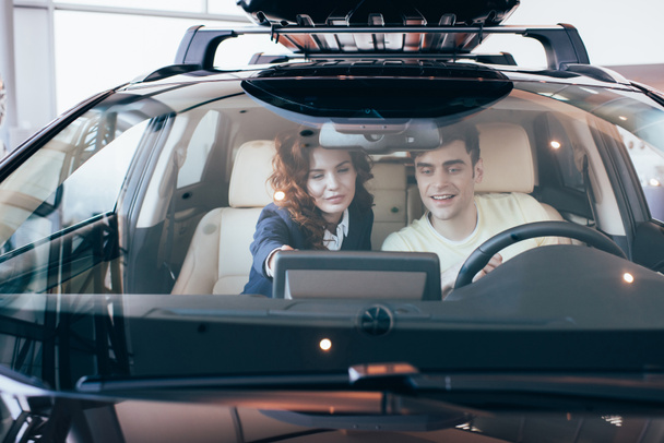 selective focus of smiling car dealer and customer sitting together in new car - Foto, Imagen