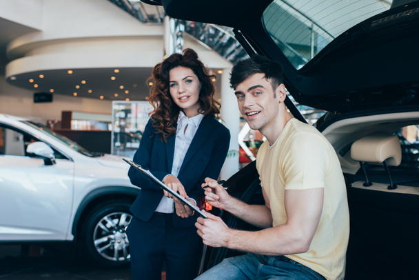 handsome man signing paper near smiling car dealer and looking at camera - Photo, Image