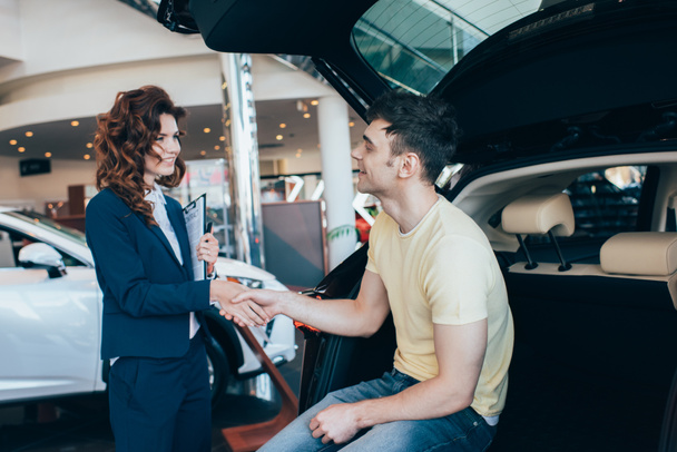 selective focus of happy man and pretty car dealer shaking hands in car showroom - Photo, Image
