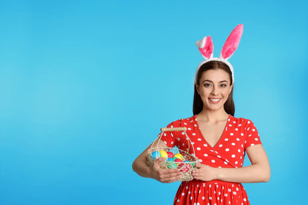 Beautiful woman in bunny ears headband holding basket with Easter eggs on color background, space for text - Photo, image