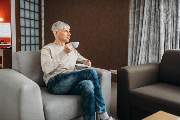 Adult man drinks coffee and reading newspaper at home. Mature male person in jeans relaxes in living room - Foto, afbeelding