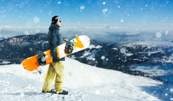 Чоловік сноубордист з дошкою стоїть на вершині засніженої гори. Сноубординг - екстремальний зимовий вид спорту. Чоловік зі сноубордом перед небезпечним схилом
 - Фото, зображення