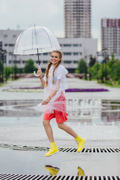 Young pretty girl with two braids in yellow boots and with transparent umbrella stands near fountain. - Photo, Image