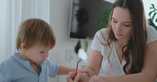 Η μαμά βοηθά δύο γιους του 2 και 4 ετών να κάνουν την προσχολική εργασία για να σχεδιάσετε μια εικόνα με μολύβια - Πλάνα, βίντεο