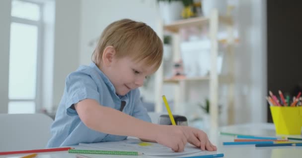 Uśmiechnięty chłopiec w niebieska koszula rysuje na papierze ołówkiem siedząc przy stole w pokoju dziennym - Materiał filmowy, wideo