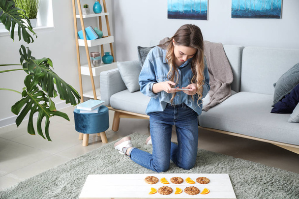 Θηλυκό τροφίμων φωτογράφος με κινητό τηλέφωνο, λαμβάνοντας εικόνα νόστιμο cookies και φέτες πορτοκαλιού στο σπίτι - Φωτογραφία, εικόνα