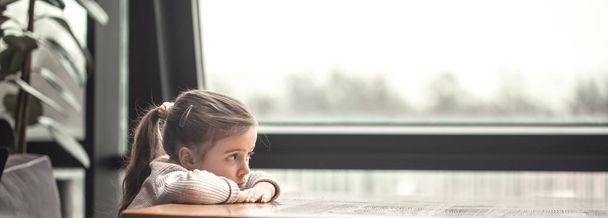 Очаровательная маленькая девочка сидит за деревянным столом у окна
. - Фото, изображение