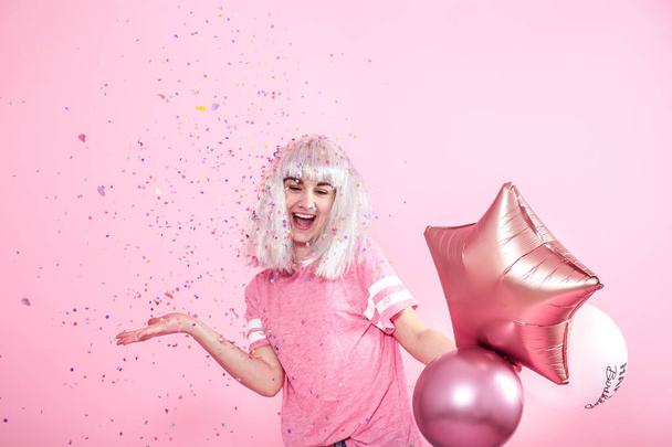 Grappig meisje met zilveren haren geeft een glimlach en emotie op roze achtergrond. Jonge vrouw of tiener meisje met ballonnen en confetti - Foto, afbeelding