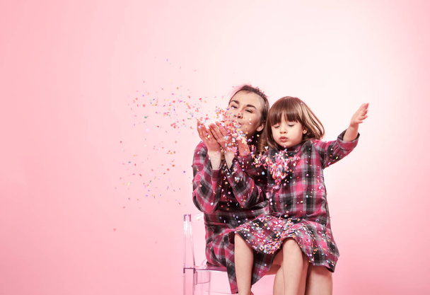 Funny mom and child sitting on a transparent stylish chairs pink background. Little girl and mother having fun with confetti - Foto, Imagem