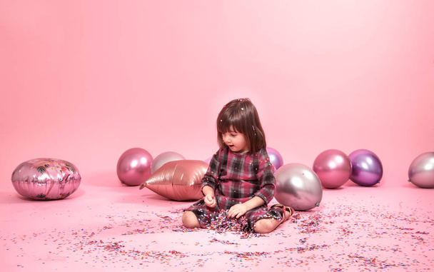 Αστείο παιδί που κάθεται σε ένα ροζ φόντο. Μικρό κορίτσι διασκεδάζοντας με μπαλόνια και κομφετί - Φωτογραφία, εικόνα