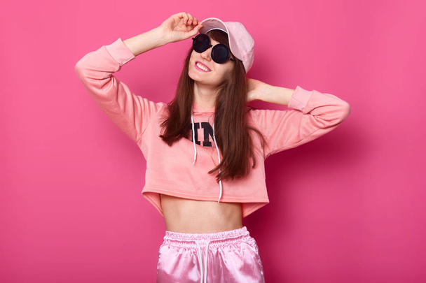 Portrait of happy smiling Caucasian brunette girl in stylish short hoodie, sweatpants and rose cap, wears black sunglasses, holds hads on hat. Poses against pink background. Teenagers concept. - Photo, Image