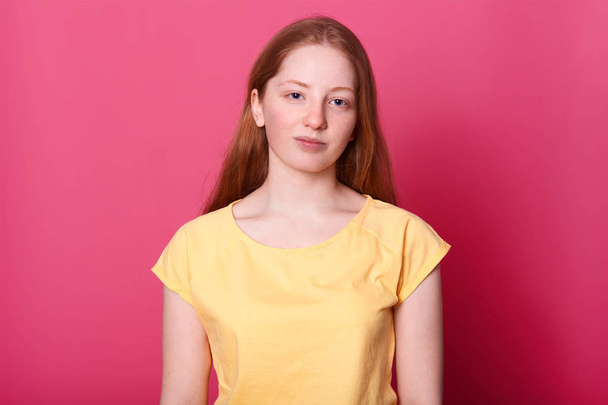 Calm schoolgirl of gentle appearance, naturally posing in studio alone, looks into camera isolated over pink background. Charming girl wears yellow t shirt, with straight brown hair. People concept. - Foto, immagini