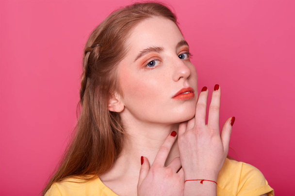 Gentle woman with bright makeup, straight hair with red shade, has manicured hand. Model in yellow shirt, keeps hands near chin, looks at camera, has mysteriously facial expression. Beauty concept. - Фото, изображение