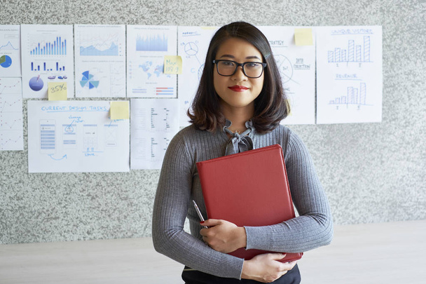 Портрет впевненої молодої бізнес-леді в окулярах, що тримає теку і стоїть в офісі з фінансовими графіками і графіками на задньому плані
 - Фото, зображення