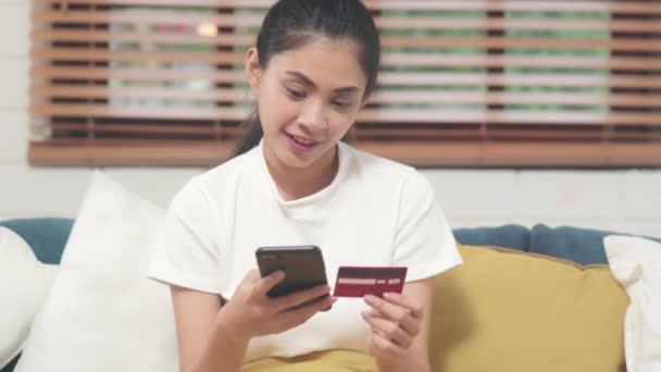 Jonge lachende Aziatische vrouw met behulp van smartphone kopen online winkelen met een creditcard terwijl liggend op de bank wanneer ontspannen in de woonkamer thuis. Lifestyle latijn en Latijns-Amerikaanse etniciteit vrouwen bij huisconcept. - Video