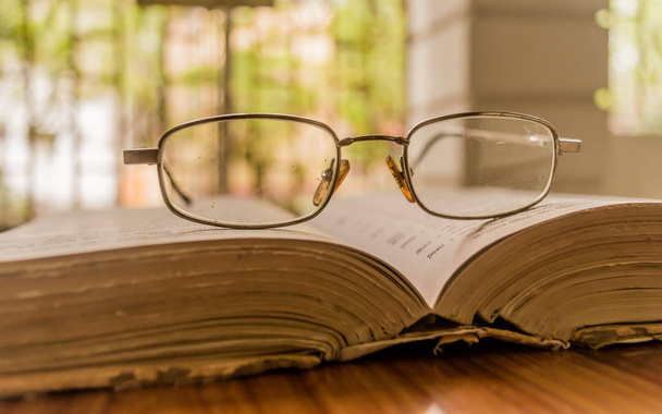 An open book on table and a specs or eyeglass on wooden table, on a warm sunny morning, side view close up. Sunday Holiday Concept. Isolated background in selective focus with shallow depth of field. - Photo, image