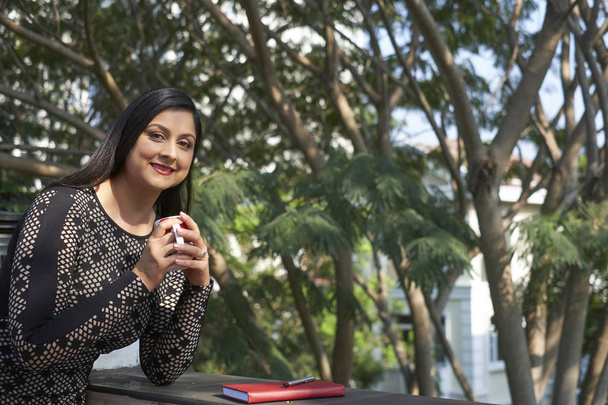 Портрет улыбающейся индийской бизнесвумен, стоящей на балконе с чашкой кофе
  - Фото, изображение