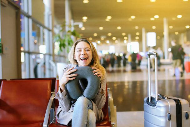 Młoda dziewczyna siedzi Lotnisko czeka sala z Poduszka pod szyję i walizka. - Zdjęcie, obraz