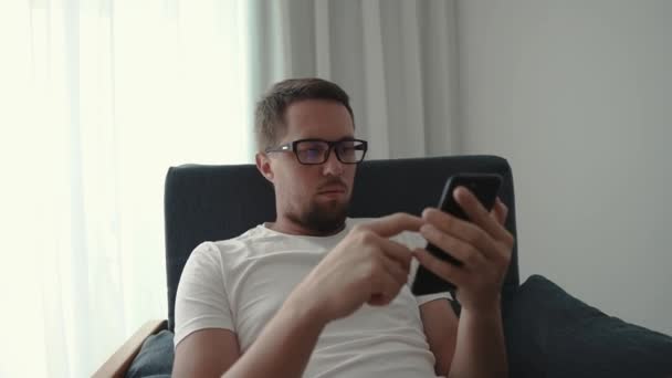 Young man with glasses is using smartphone in room in daytime, close-up view - Záběry, video