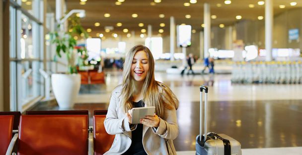 Молодая красивая женщина сидит в зале аэропорта с планшетом и чемоданом, используя Интернет
. - Фото, изображение