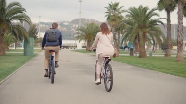 Man and woman are riding bikes in city park in cloudy day in summer, back view - Footage, Video