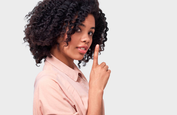 Confidential African American young woman dressed in pink shirt holding index finger on lips, asking to keep silence over white wall. Beautiful dark skinned female looks to the camera asks to be quiet - Фото, изображение