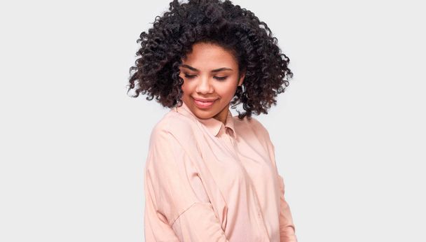 Dreamy dark-skinned woman smiling broadly with closed eyes, wearing beige shirt, enjoy good time, posing against white studio wall. People, success, emotions and happiness concept - Photo, Image
