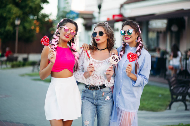 Τρία κορίτσια που ποζάρουν στο δρόμο εκμετάλλευση γλυκά καραμέλες σε stick. - Φωτογραφία, εικόνα