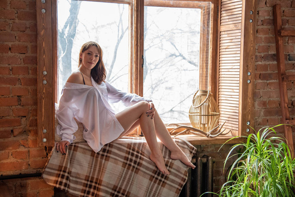 Beautiful girl in a white shirt is sitting on the windowsill in  - Fotó, kép