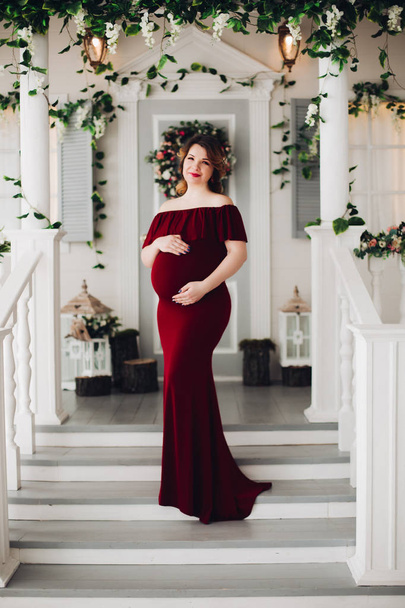 Charming pregnant woman in marsala dress posing on steps of house. - Photo, image